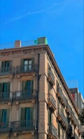 A multi-story residential building with classic architectural design featuring multiple balconies with wrought iron railings. The structure has a beige facade with green window shutters and is set against a clear blue sky.