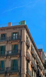 A multi-story residential building with classic architectural design featuring multiple balconies with wrought iron railings. The structure has a beige facade with green window shutters and is set against a clear blue sky.
