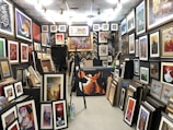 A gallery space filled with numerous framed artworks covering the walls and floor. The artworks range from abstract pieces to portraits and calligraphy, displayed in various sizes and frames. The room is well-lit with multiple ceiling lights, and there are some empty frames leaning against the walls.
