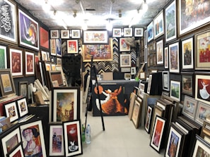 A gallery space filled with numerous framed artworks covering the walls and floor. The artworks range from abstract pieces to portraits and calligraphy, displayed in various sizes and frames. The room is well-lit with multiple ceiling lights, and there are some empty frames leaning against the walls.