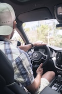 A person is seated in the driver's seat of a car, wearing a plaid shirt and a cap. The steering wheel features the Volvo logo. The interior of the car appears modern with a visible dashboard and console. The view outside through the windshield shows trees and a clear sky.