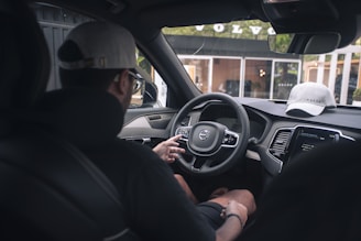 A person is seated in the driver's seat of a car, interacting with the steering wheel and dashboard controls. A cap is placed on the dashboard, and the name of the car brand is visible on a nearby building through the window.