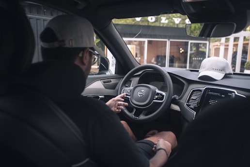 A person is seated in the driver's seat of a car, interacting with the steering wheel and dashboard controls. A cap is placed on the dashboard, and the name of the car brand is visible on a nearby building through the window.