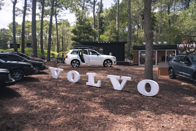 A white Volvo SUV with its doors open is parked among trees in a forested area. Large white letters on the ground spell out 'VOLVO' in front of the vehicle. The background includes other parked cars and a black shipping container with the Volvo logo. A bicycle is mounted on top of one of the vehicles to the right.