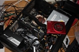 An open computer case with various components visible, including a Corsair VS650 power supply, a graphics card, and a Sound Blaster card. There are wires and cables loosely organized within and around the case. The setup is on a wooden surface, and in the background, numerous tangled cables are also visible.