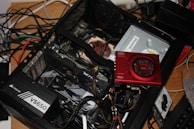An open computer case with various components visible, including a Corsair VS650 power supply, a graphics card, and a Sound Blaster card. There are wires and cables loosely organized within and around the case. The setup is on a wooden surface, and in the background, numerous tangled cables are also visible.