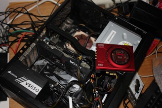 An open computer case with various components visible, including a Corsair VS650 power supply, a graphics card, and a Sound Blaster card. There are wires and cables loosely organized within and around the case. The setup is on a wooden surface, and in the background, numerous tangled cables are also visible.