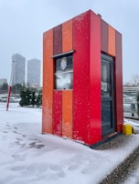A brightly colored booth stands in a snowy environment. The booth features vertical stripes in red and orange shades, with snow accumulating on its surface. In the background, modern high-rise buildings and evergreen trees are visible, and snow covers the ground, with footprints marking the path around the booth.