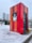 A brightly colored booth stands in a snowy environment. The booth features vertical stripes in red and orange shades, with snow accumulating on its surface. In the background, modern high-rise buildings and evergreen trees are visible, and snow covers the ground, with footprints marking the path around the booth.