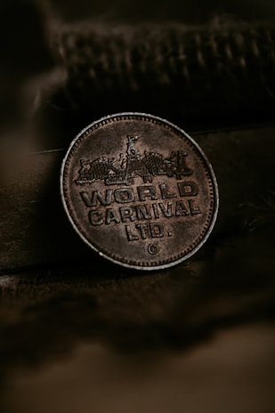 A close-up of a vintage, metallic token or coin embossed with the words 'World Carnival Ltd.' It features intricate designs depicting a carnival scene with tents and a ferris wheel. The backdrop has a rustic texture with an earthy tone, enhancing the antique feel of the coin.