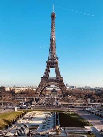 A clear view of the Eiffel Tower in Paris, set against a bright blue sky. The foreground includes landscaped gardens with pathways and fountains, while the background features Parisian architecture and cityscape.
