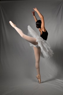 A ballerina is gracefully performing a high leg extension while balancing on pointe shoes. She wears a black leotard and a white tutu, displaying poise and elegance.