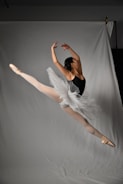 A ballet dancer is captured mid-leap, demonstrating an elegant and dynamic pose. The dancer wears a white tutu and a black leotard, highlighting the athleticism and grace of the movement. The background is a neutral gray, which accentuates the dancer's form and the fluidity of the gesture.