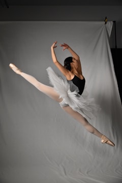 A ballet dancer is captured mid-leap, demonstrating an elegant and dynamic pose. The dancer wears a white tutu and a black leotard, highlighting the athleticism and grace of the movement. The background is a neutral gray, which accentuates the dancer's form and the fluidity of the gesture.