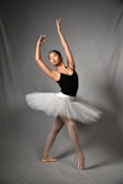 A dancer poses gracefully in a ballet position. She wears a black leotard paired with a white tutu, her arms elegantly extended above her head. The background is a plain, soft gray, and her feet are in pointe shoes, reflecting the discipline and artistry of ballet.