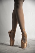 A pair of feet in ballet pointe shoes, elegantly posed on a neutral backdrop. The shoes and legs are gracefully positioned, showcasing poise and alignment typical of ballet dancers.