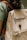 A person is wearing a green shirt and carrying a beige backpack with a brown strap. The backpack has the text 'Kawê Rodrigues Fotografia' embroidered on it and a round button with a logo. The scene has a casual and natural feel.