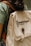 A person is wearing a green shirt and carrying a beige backpack with a brown strap. The backpack has the text 'Kawê Rodrigues Fotografia' embroidered on it and a round button with a logo. The scene has a casual and natural feel.