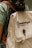 A person is wearing a green shirt and carrying a beige backpack with a brown strap. The backpack has the text 'Kawê Rodrigues Fotografia' embroidered on it and a round button with a logo. The scene has a casual and natural feel.