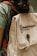 A person is wearing a green shirt and carrying a beige backpack with a brown strap. The backpack has the text 'Kawê Rodrigues Fotografia' embroidered on it and a round button with a logo. The scene has a casual and natural feel.