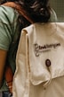 A person is wearing a green shirt and carrying a beige backpack with a brown strap. The backpack has the text 'Kaw&ecirc; Rodrigues Fotografia' embroidered on it and a round button with a logo. The scene has a casual and natural feel.