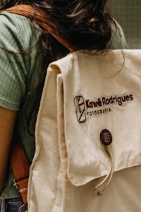 A person is wearing a green shirt and carrying a beige backpack with a brown strap. The backpack has the text 'Kaw&ecirc; Rodrigues Fotografia' embroidered on it and a round button with a logo. The scene has a casual and natural feel.