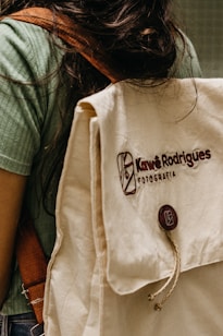 A person is wearing a green shirt and carrying a beige backpack with a brown strap. The backpack has the text 'Kaw&ecirc; Rodrigues Fotografia' embroidered on it and a round button with a logo. The scene has a casual and natural feel.