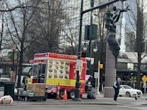 A vibrant red food truck is parked on the side of a city street, featuring a large menu depicting various dishes. Nearby, a person stands beside the truck, and a sculpture of a human figure holding a traffic light is visible. Leafless trees and urban buildings provide a backdrop, along with street signs and a white parked car.