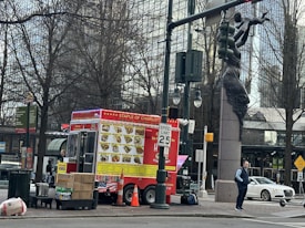 A vibrant red food truck is parked on the side of a city street, featuring a large menu depicting various dishes. Nearby, a person stands beside the truck, and a sculpture of a human figure holding a traffic light is visible. Leafless trees and urban buildings provide a backdrop, along with street signs and a white parked car.