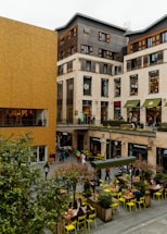 A bustling urban area featuring modern commercial buildings with a mix of wood and beige stone facades. The scene includes multiple shops and restaurants such as Starbucks and McDonald's, with people walking and sitting at outdoor tables. Bright yellow chairs and small trees in planter boxes add a pop of color to the otherwise neutral-toned setting.