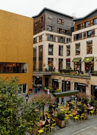 A bustling urban area featuring modern commercial buildings with a mix of wood and beige stone facades. The scene includes multiple shops and restaurants such as Starbucks and McDonald's, with people walking and sitting at outdoor tables. Bright yellow chairs and small trees in planter boxes add a pop of color to the otherwise neutral-toned setting.