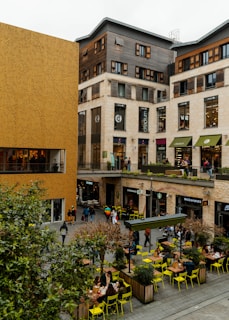 A bustling urban area featuring modern commercial buildings with a mix of wood and beige stone facades. The scene includes multiple shops and restaurants such as Starbucks and McDonald's, with people walking and sitting at outdoor tables. Bright yellow chairs and small trees in planter boxes add a pop of color to the otherwise neutral-toned setting.