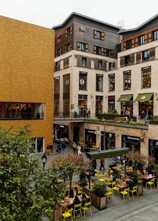 A bustling urban area featuring modern commercial buildings with a mix of wood and beige stone facades. The scene includes multiple shops and restaurants such as Starbucks and McDonald's, with people walking and sitting at outdoor tables. Bright yellow chairs and small trees in planter boxes add a pop of color to the otherwise neutral-toned setting.