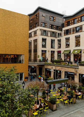 A bustling urban area featuring modern commercial buildings with a mix of wood and beige stone facades. The scene includes multiple shops and restaurants such as Starbucks and McDonald's, with people walking and sitting at outdoor tables. Bright yellow chairs and small trees in planter boxes add a pop of color to the otherwise neutral-toned setting.