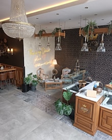 The interior of a cozy, upscale café or patisserie with elegant décor. Hanging from the ceiling is a large, ornate chandelier. The walls feature a mix of patterned wallpaper and a modern art piece with neon lettering. The space includes stylish, vintage-style chairs and a table with a gramophone atop it. There are also large glass display cases and potted plants adding greenery to the space.