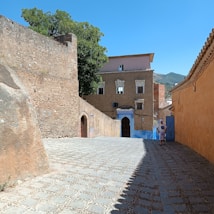 A cobblestone street flanked by textured, earthy-colored walls leads to a building with blue accents. The alleyway is framed by tall, rustic walls, and there's a person walking in the shade, dressed in light clothing. In the background, trees and a mountainous landscape are visible under a bright blue sky.
