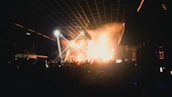 A crowded nightclub with a lively atmosphere. Bright lights and smoke fill the space as people raise their hands in excitement. The stage lights illuminate the area, casting vibrant colors and shadows across the crowd.