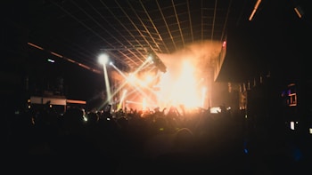 A crowded nightclub with a lively atmosphere. Bright lights and smoke fill the space as people raise their hands in excitement. The stage lights illuminate the area, casting vibrant colors and shadows across the crowd.