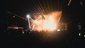 A crowded nightclub with a lively atmosphere. Bright lights and smoke fill the space as people raise their hands in excitement. The stage lights illuminate the area, casting vibrant colors and shadows across the crowd.