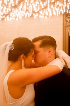 A bride and groom share an intimate moment, touching foreheads with their eyes closed. The bride is wearing a white dress with a veil, adorned with a sparkling hair accessory, and long dangling earrings. The groom is in a dark suit. The background features a warm, ambient lighting with numerous small lights, creating a romantic atmosphere.