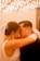 A bride and groom share an intimate moment, touching foreheads with their eyes closed. The bride is wearing a white dress with a veil, adorned with a sparkling hair accessory, and long dangling earrings. The groom is in a dark suit. The background features a warm, ambient lighting with numerous small lights, creating a romantic atmosphere.