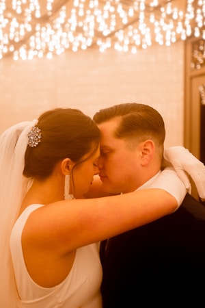 A bride and groom share an intimate moment, touching foreheads with their eyes closed. The bride is wearing a white dress with a veil, adorned with a sparkling hair accessory, and long dangling earrings. The groom is in a dark suit. The background features a warm, ambient lighting with numerous small lights, creating a romantic atmosphere.