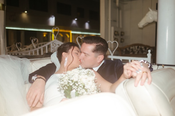 A couple is sharing a passionate kiss while seated on a white cushioned bench. The woman is dressed in a white gown, accessorized with large earrings, and is holding a bouquet of white flowers. The man is dressed in a dark suit and their hands are intertwined. The background features heart-shaped decorations and a horse figure, suggesting a romantic setting.