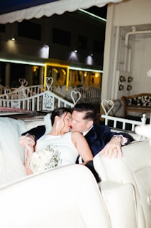 A couple is sitting closely together on a white couch, sharing an intimate moment. The woman is wearing a white dress and holding a bouquet of white flowers. The man is dressed in a dark suit. The setting appears to be an elegant outdoor area with white furniture and decorative heart-shaped metalwork.
