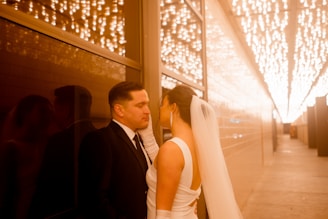 A couple stands closely together in a warmly lit hallway, with the man wearing a black suit and the woman in a white dress and veil. String lights hang above, creating a romantic ambiance with their reflection visible on the glass wall next to them.