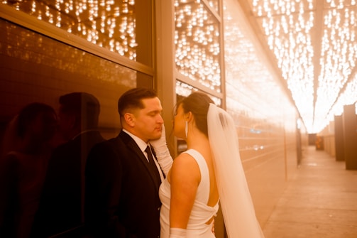 A couple stands closely together in a warmly lit hallway, with the man wearing a black suit and the woman in a white dress and veil. String lights hang above, creating a romantic ambiance with their reflection visible on the glass wall next to them.