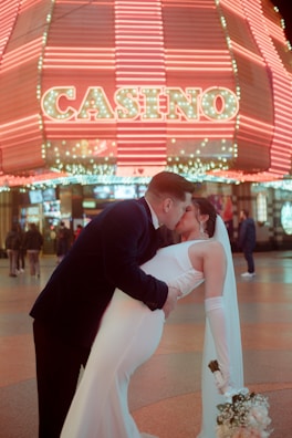 A couple is captured in a romantic pose, with the man dipping the woman for a kiss. They are dressed in formal attire, as the woman holds a bouquet. In the background, a brightly lit casino sign with neon lights creates a vibrant atmosphere. Other people are seen walking in the background, adding life to the scene.