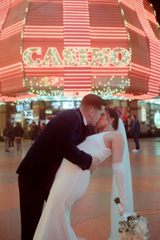 A couple is captured in a romantic pose, with the man dipping the woman for a kiss. They are dressed in formal attire, as the woman holds a bouquet. In the background, a brightly lit casino sign with neon lights creates a vibrant atmosphere. Other people are seen walking in the background, adding life to the scene.