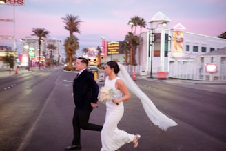 A couple is crossing a wide street with palm trees lining the sides, holding hands. The bride is wearing a white gown with a veil flowing behind her, while the groom is in a dark suit. The background features bright neon signs and modern buildings, indicating an urban setting.