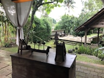 A tranquil outdoor scene featuring a wooden structure with various sculptures placed on top. There is lush greenery all around, including bamboo and other plants. The setting appears to be a serene garden with stone pathways, a small temple-like structure, and a traditional gazebo. The space is partially shaded by a white fabric curtain.